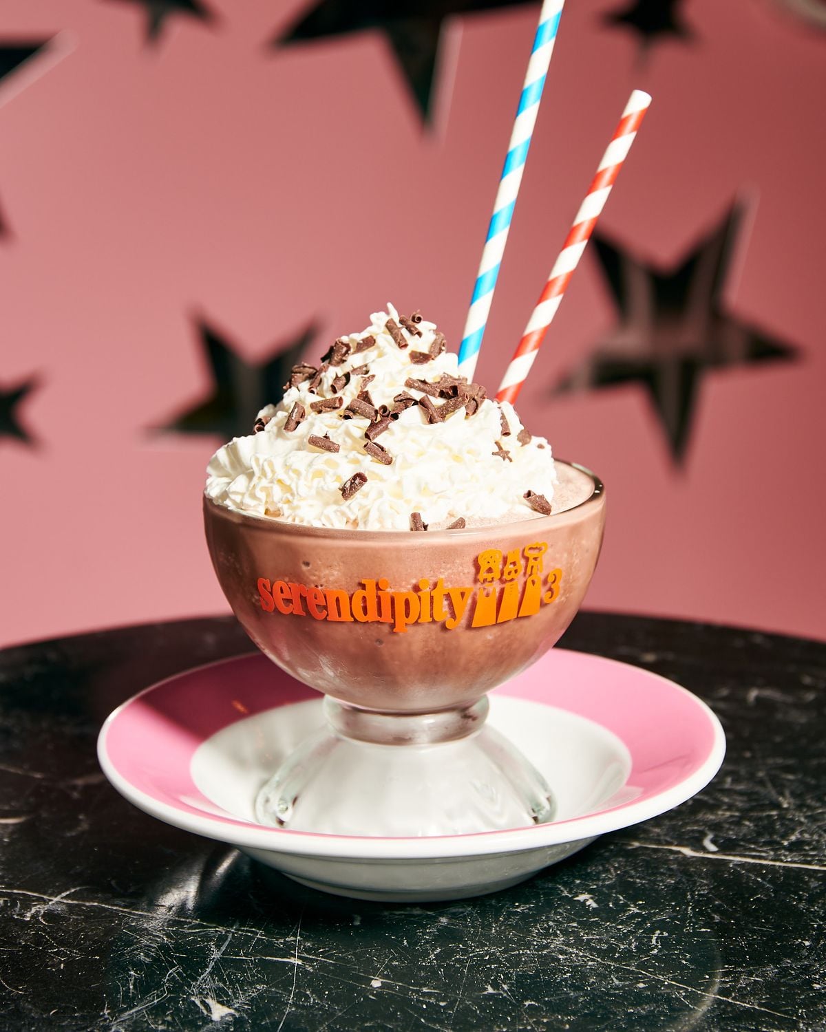 A glass cup labeled "serendipity 3" filled with a chocolate dessert topped with whipped cream and chocolate shavings, served on a pink and white plate with two striped straws, against a pink background with black stars.
