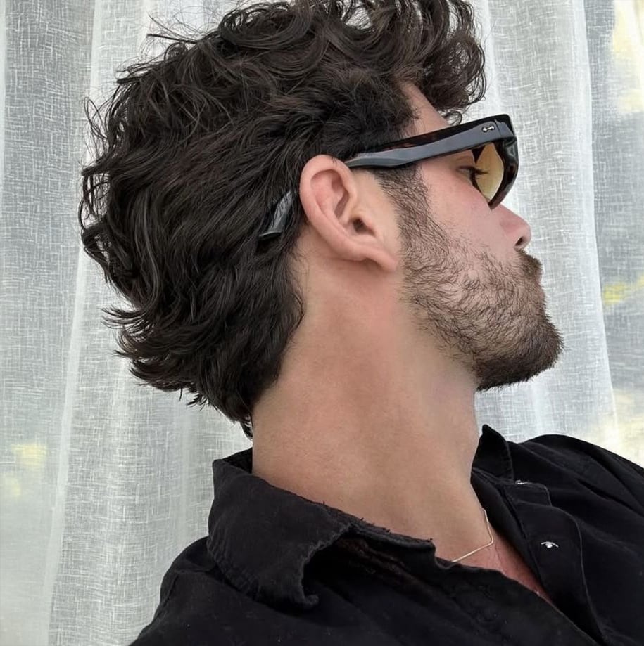 A person with wavy dark hair and a short beard is wearing black sunglasses and a black shirt, looking to the side against a sheer white curtain backdrop.