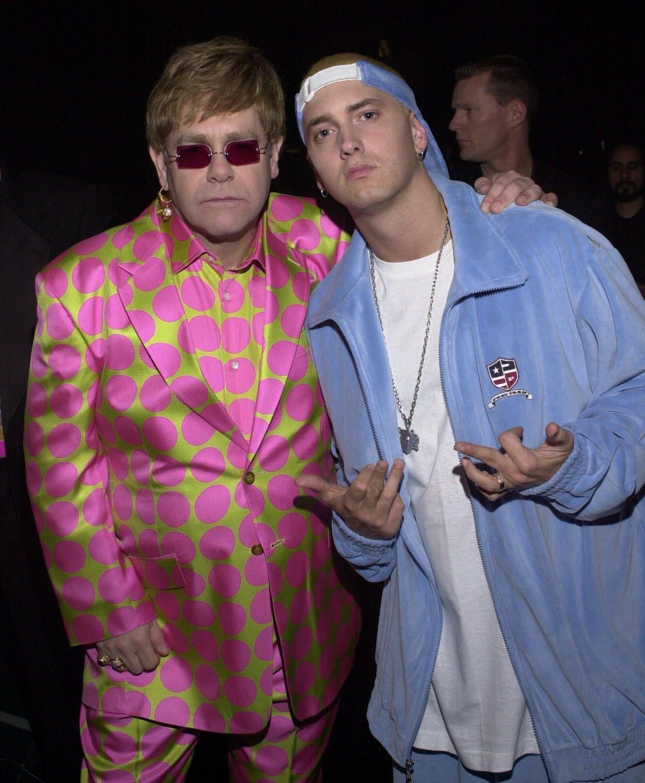 Two men pose together. One wears a bright pink suit with green polka dots and red sunglasses. The other wears a light blue tracksuit, a white t-shirt, and a backwards cap, making hand gestures. They stand against a dark background.