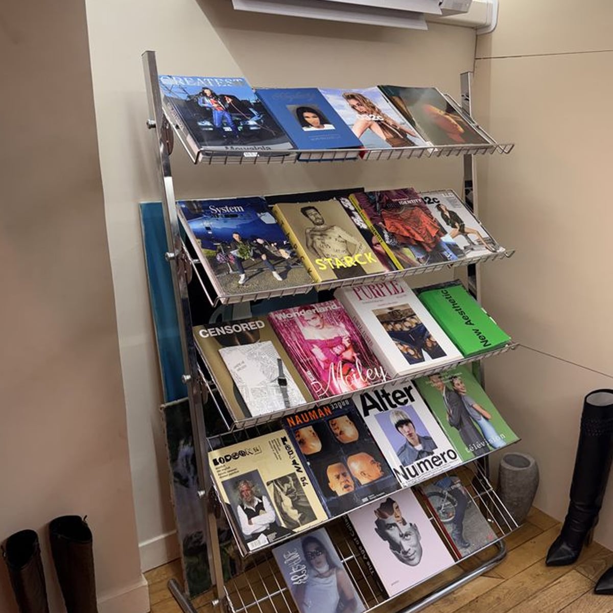 A metal magazine rack displays various colorful magazines and books on three shelves, arranged neatly. The rack stands on a wooden floor next to a pair of black boots and a gray cylindrical object.