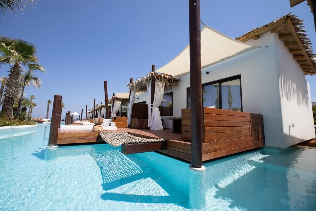 A modern white villa with wooden accents sits beside a clear blue pool, featuring private deck areas with outdoor beds and hammocks under a sunny, clear sky surrounded by palm trees.
