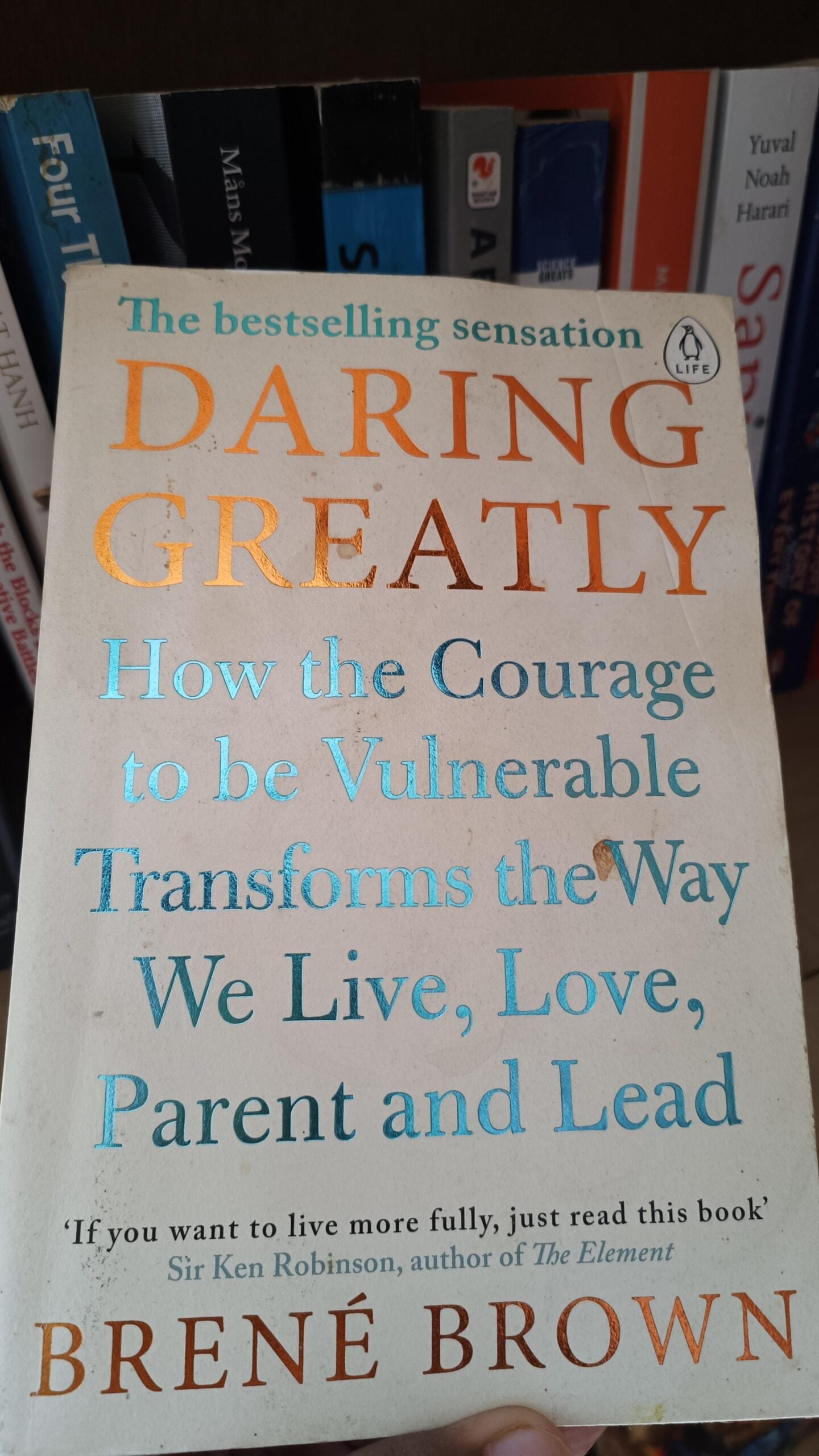 A close-up of the book "Daring Greatly" by Brené Brown, showing the cover text and part of a bookshelf with other books in the background.