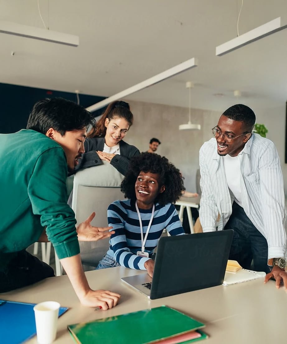 Four people gather around a desk, smiling and talking, with one person sitting and using a laptop while the others stand nearby. They appear to be collaborating in a modern office setting.