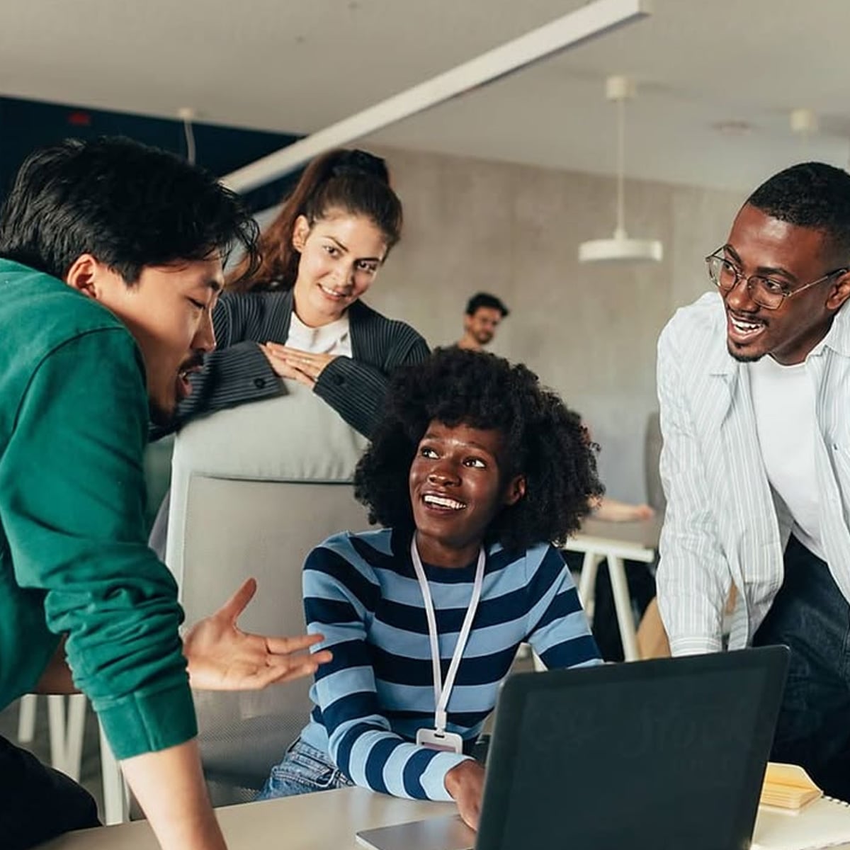 Four young adults, three standing and one sitting at a desk with a laptop, engage in a lively discussion in a modern office setting. They are smiling and appear to be collaborating on a project.