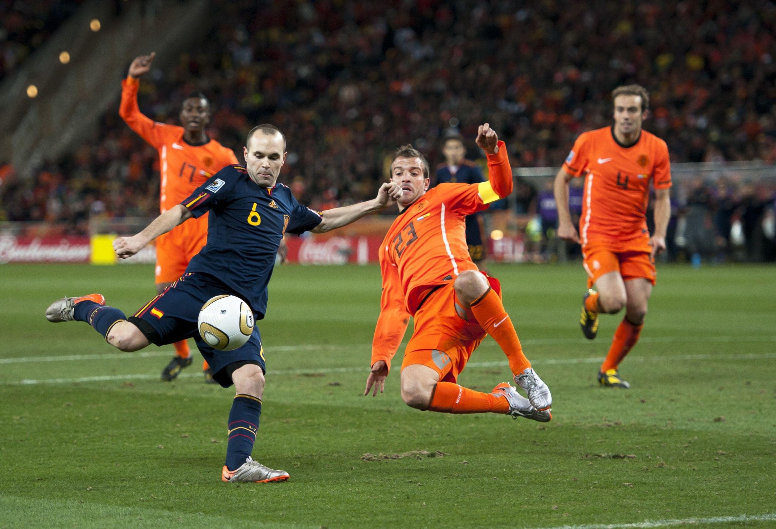 A soccer player in a dark blue uniform kicks the ball as an opponent in an orange uniform slides to block him. Two other players in orange and a crowd in the stadium are visible in the background.