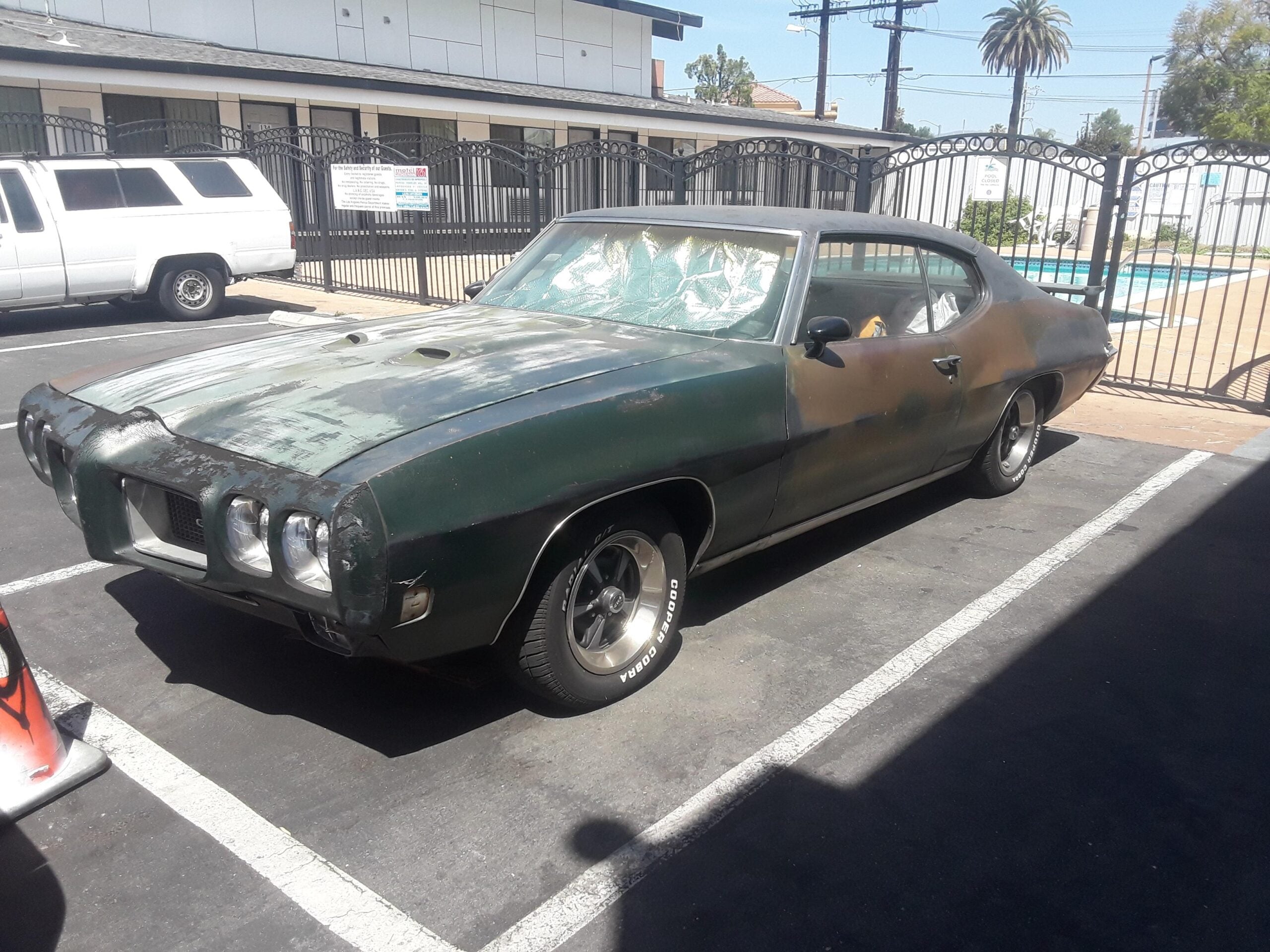 A weathered, dark green vintage muscle car with faded paint is parked in a lot. The windshield is covered with a reflective sunshade, and there is a white SUV and a gated pool area in the background.