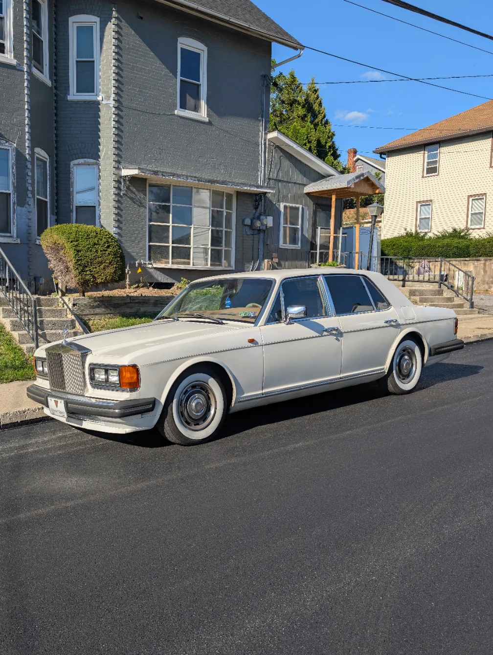 A vintage white luxury car, likely a 1980s Rolls-Royce, is parked on a freshly paved residential street beside gray and tan houses under a clear blue sky.