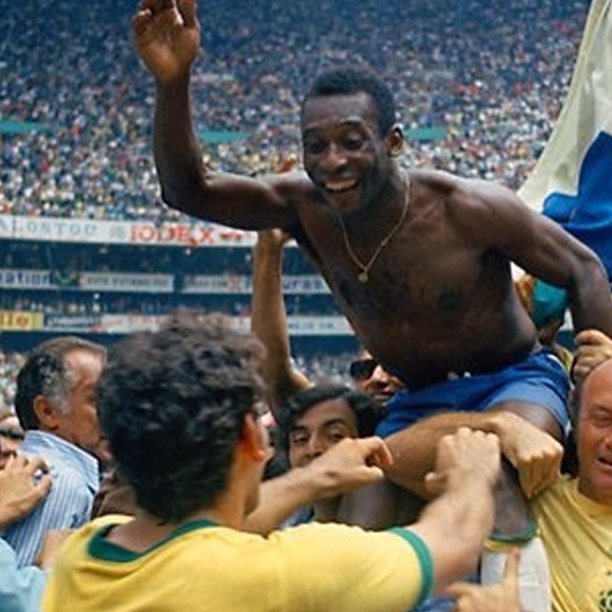 A soccer player in blue shorts is lifted and celebrated by a crowd in a stadium filled with spectators. He is smiling, raising an arm, and appears joyful amid the excitement.