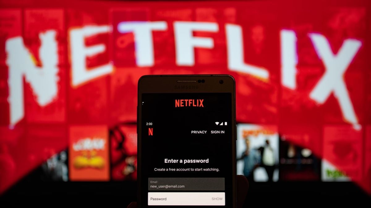 A smartphone displays the Netflix login screen, asking for a password, with a large blurry Netflix logo in bold white letters on a bright red background visible on a screen behind it.
