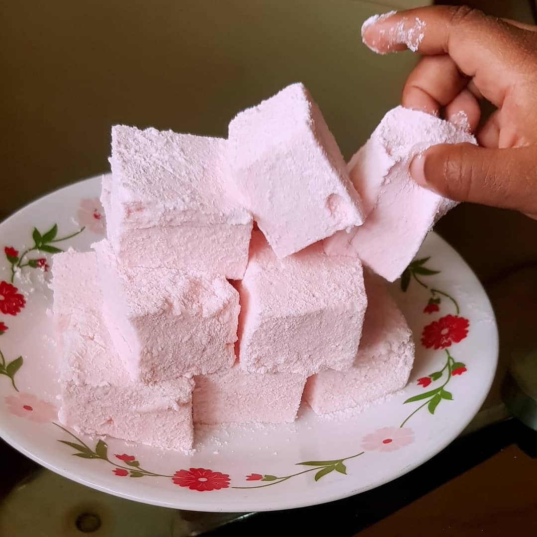 A plate with a stack of pink, powdered sugar-coated cubes, resembling marshmallows. A hand is reaching to pick up one piece. The plate has a floral pattern along the rim.