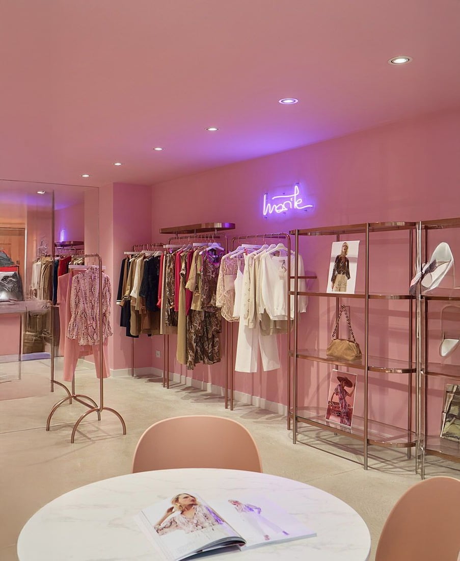 Chic boutique with pink walls and ceiling, featuring racks of stylish clothing, shelves displaying bags and shoes, and a round table with chairs and an open fashion magazine. Neon sign on the wall glows purple.