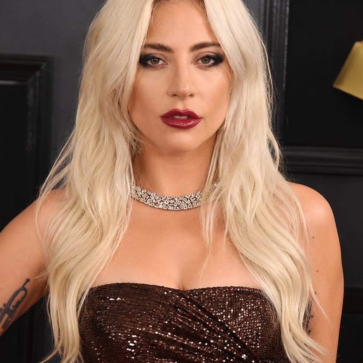 A woman with long platinum blonde hair wears a sparkling, strapless dark dress and a diamond necklace. She has bold dark eye makeup and dark red lipstick, standing in front of a black background.
