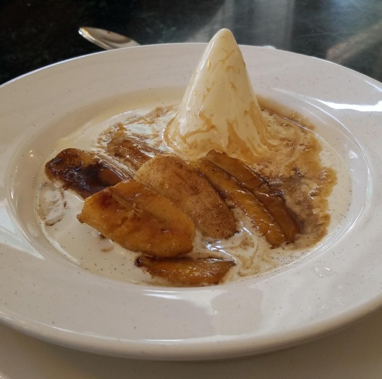 A white plate holds caramelized banana slices in sauce, served with a cone-shaped scoop of vanilla ice cream, melting slightly. A metal spoon is visible in the background.