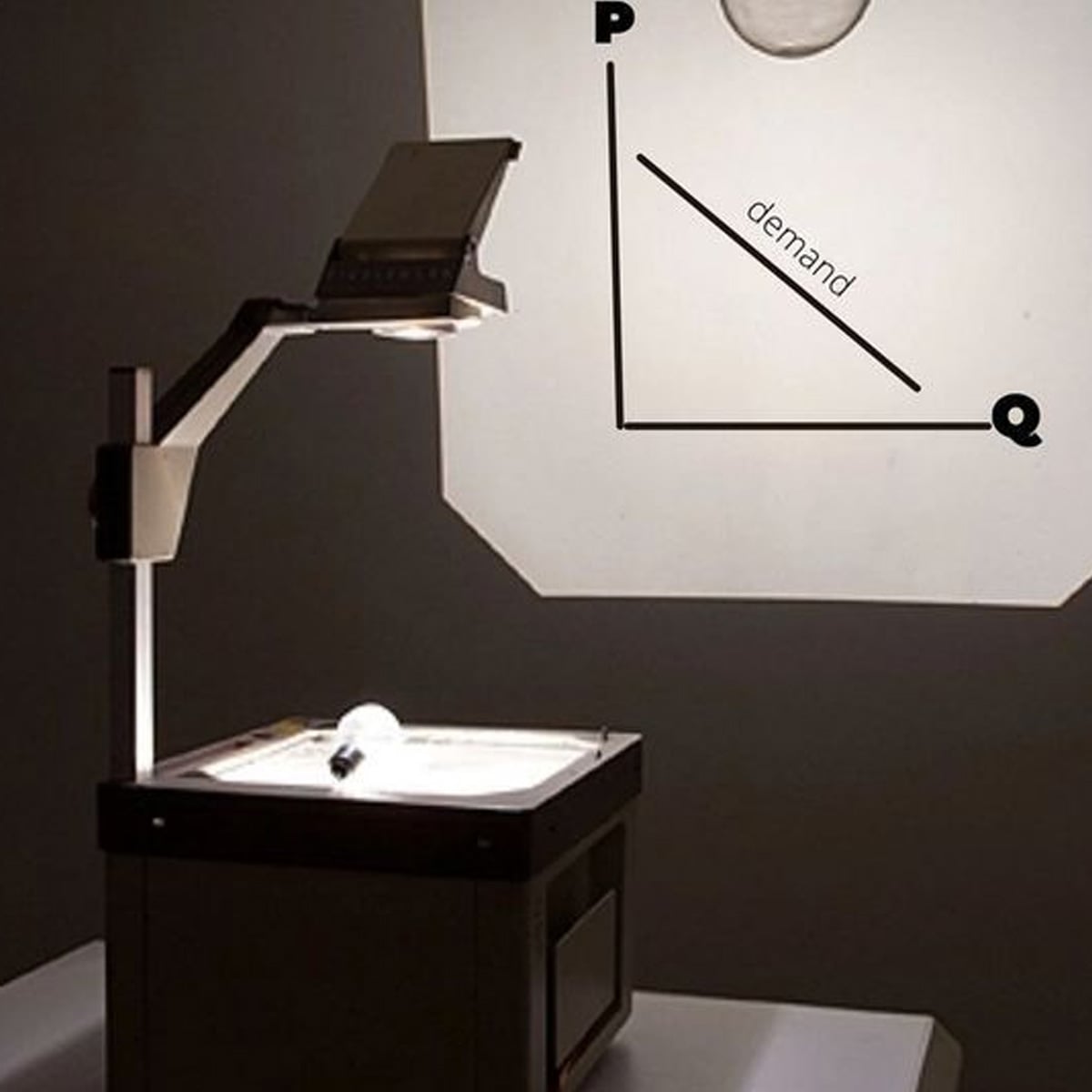 An overhead projector displays a simple demand curve graph labeled "P" for price and "Q" for quantity on a wall, with the word "demand" along a downward-sloping line.