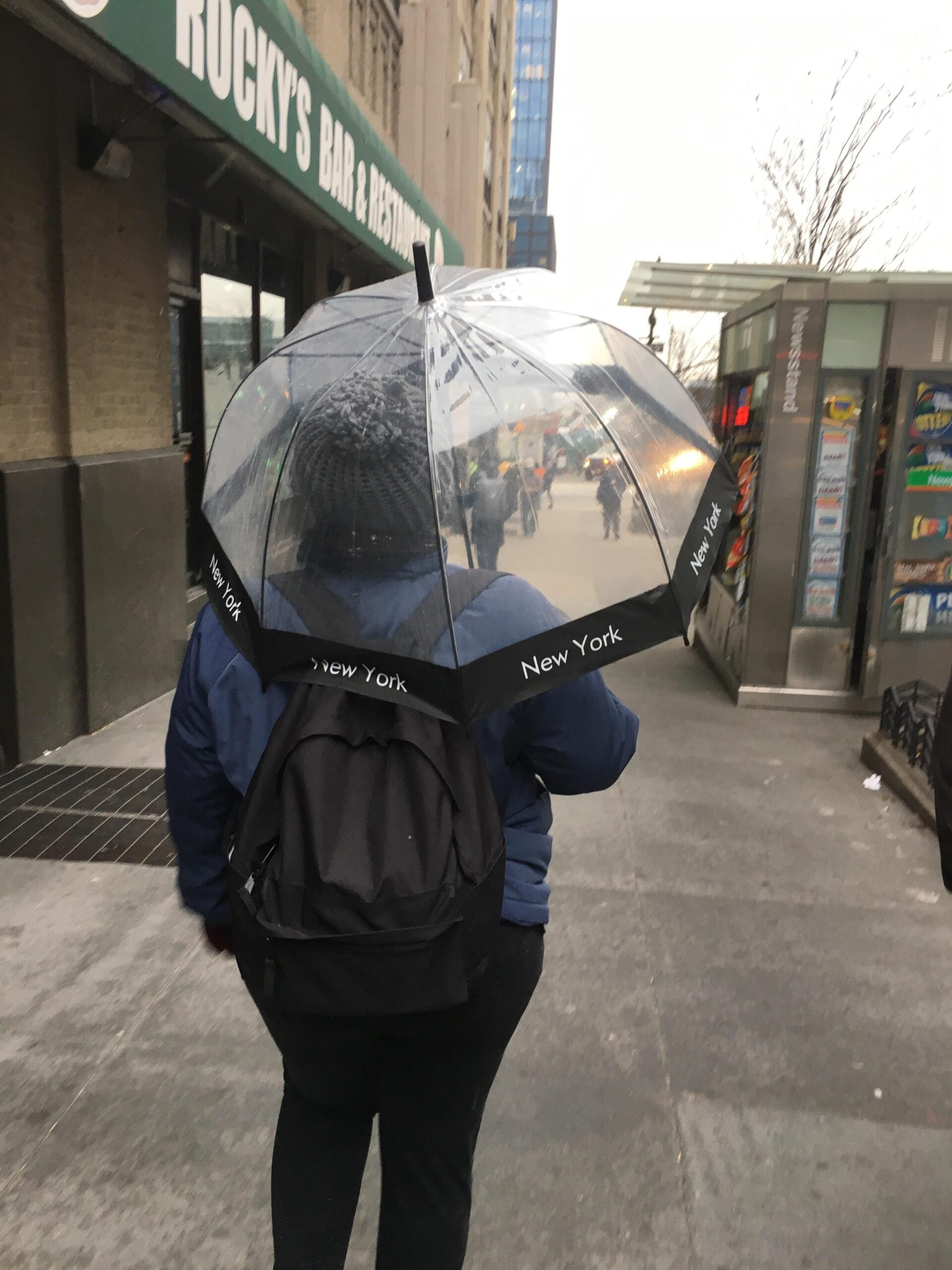 A person with a black backpack walks down a city sidewalk under a transparent umbrella labeled “New York.” Buildings, a newsstand, and pedestrians are visible in the background.