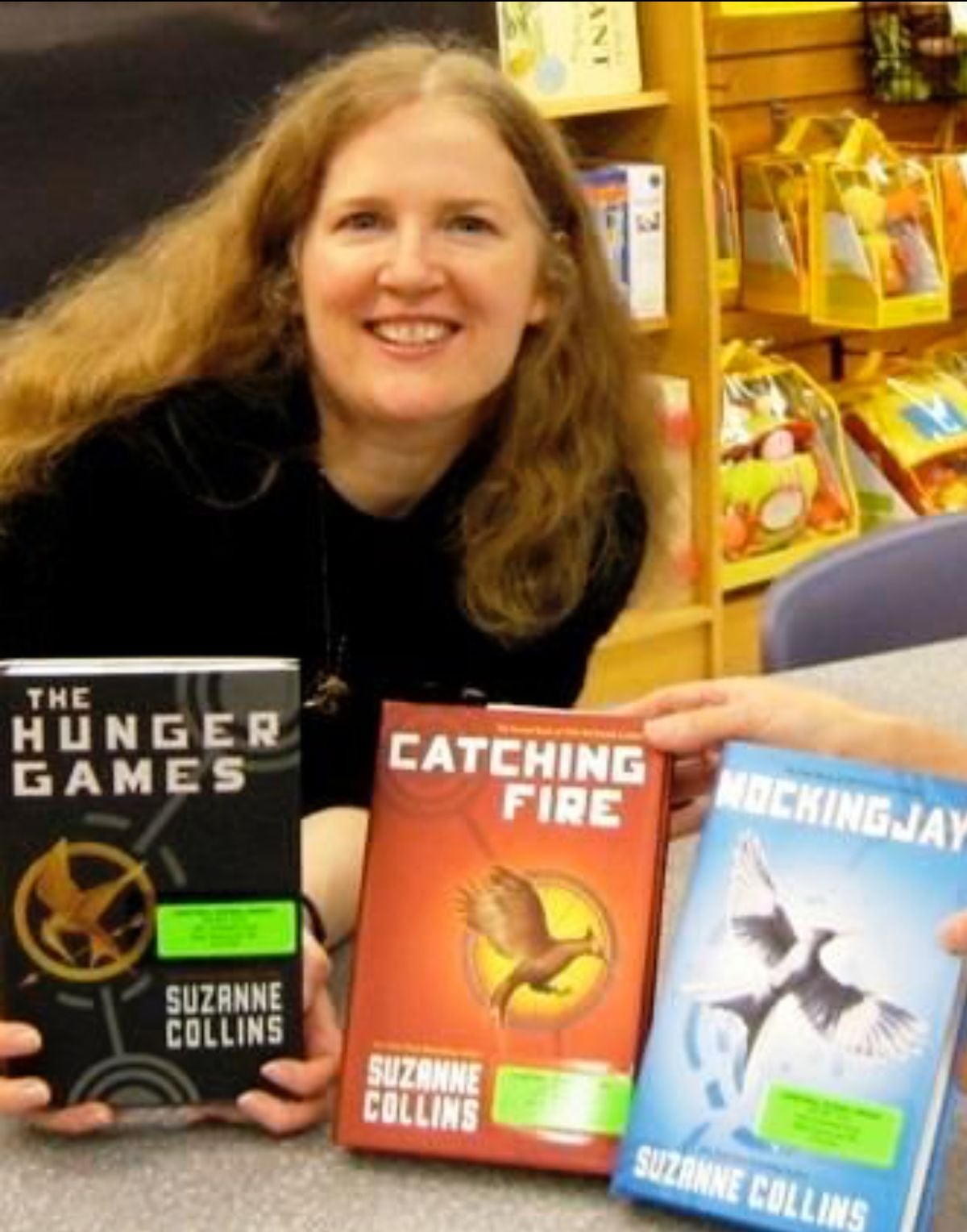 A smiling woman with long hair sits at a table holding three books from The Hunger Games series: "The Hunger Games," "Catching Fire," and "Mockingjay," all by Suzanne Collins.
