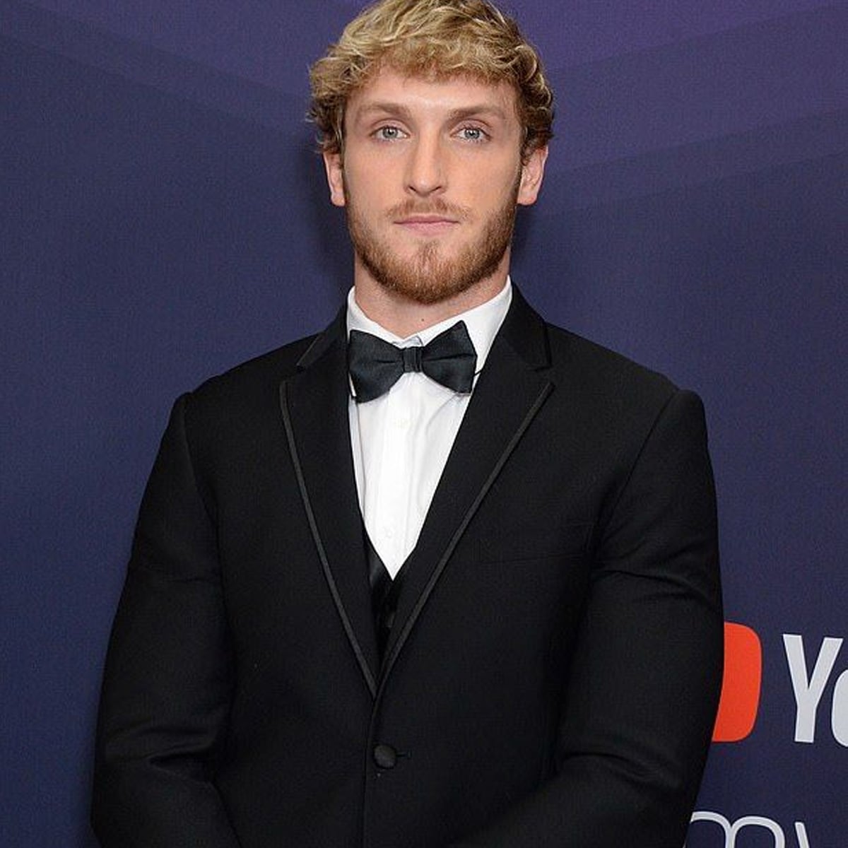 A man with short, curly blond hair and a trimmed beard wears a black tuxedo with a bow tie, standing in front of a dark blue backdrop with partial YouTube and red play button logos.