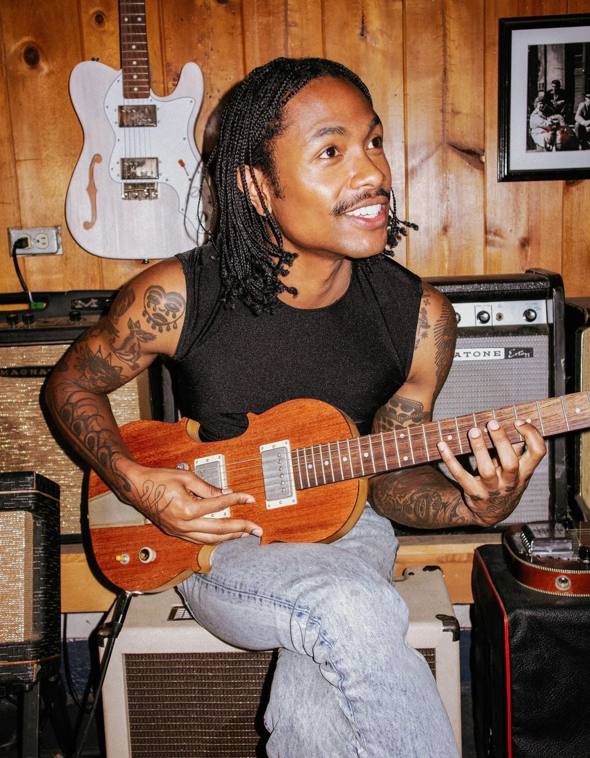 A person with braided hair and tattoos sits on an amplifier, playing a brown electric guitar. They wear a sleeveless black top and light jeans. Musical equipment and a guitar hang on a wooden wall behind them.