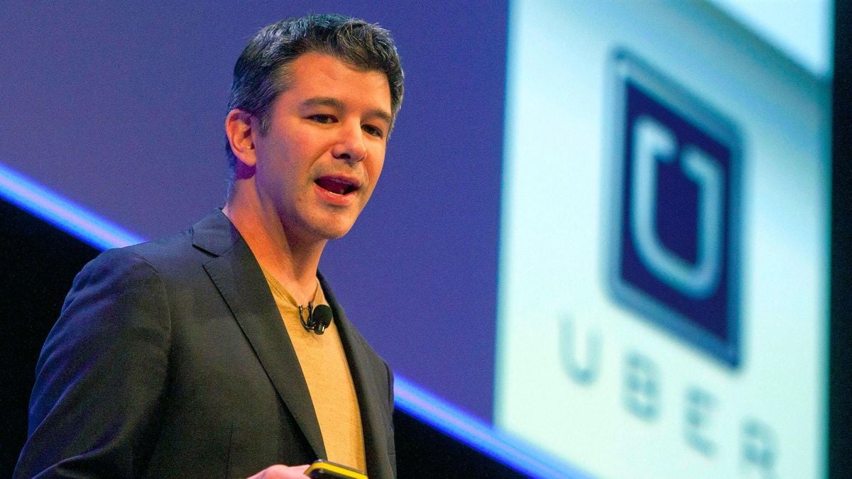A man in a dark blazer speaks on stage, holding a small device. Behind him is a large screen displaying the Uber logo.