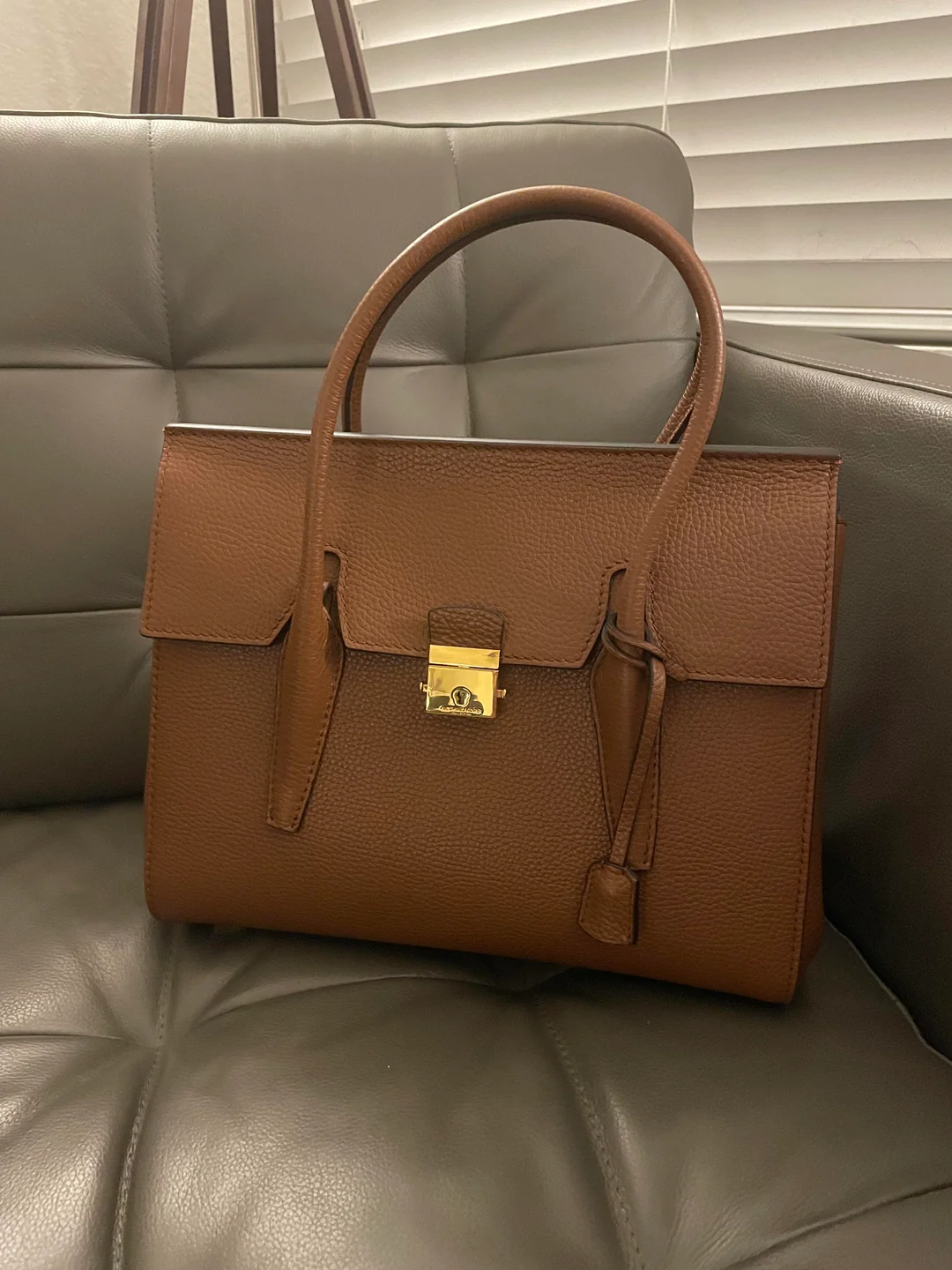 A brown leather handbag with a structured shape and a gold clasp sits on a gray cushioned sofa in front of white window blinds.