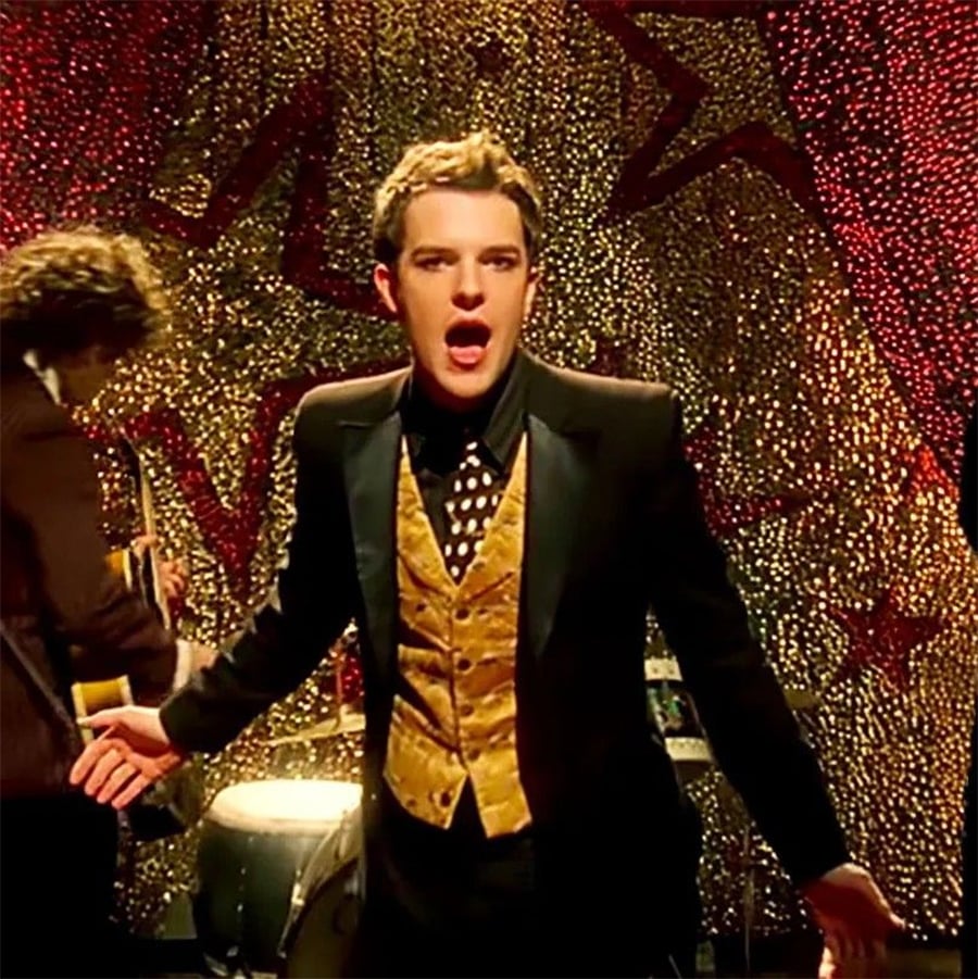 A man in a black suit with a gold vest and polka dot tie sings passionately in front of a sparkling gold backdrop with red stars, while another musician plays guitar beside him.