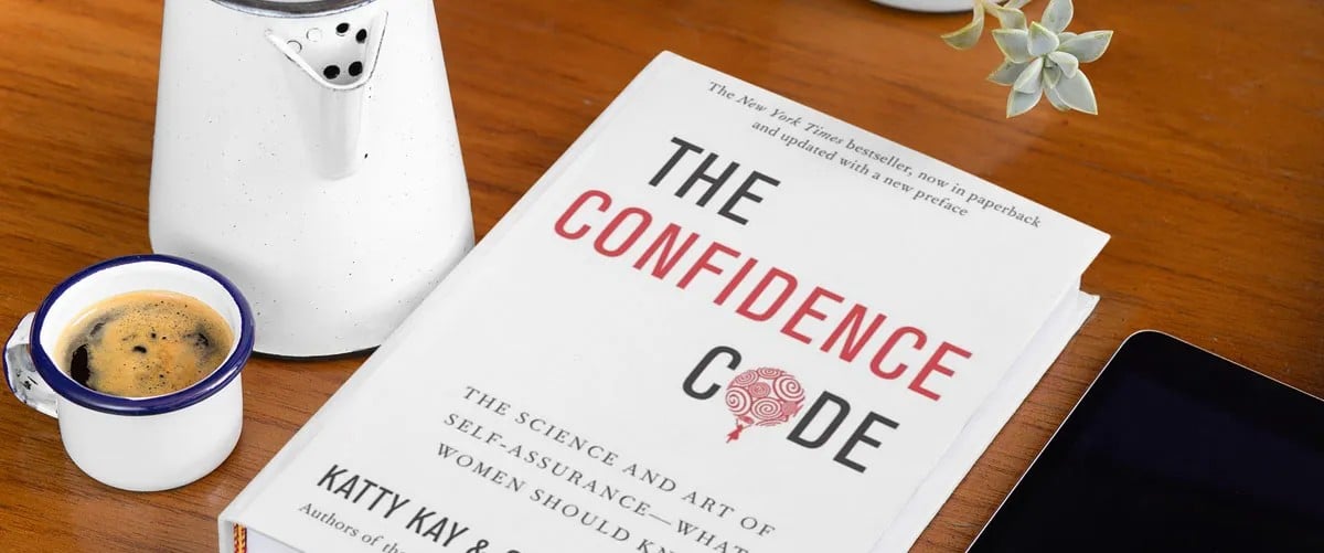A copy of “The Confidence Code” by Katty Kay and Claire Shipman rests on a wooden table next to a cup of coffee, a white enamel coffee pot, a small potted succulent, and a tablet.