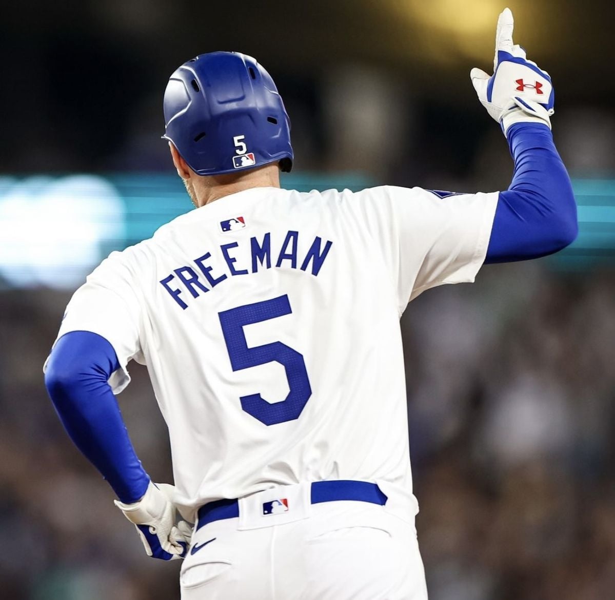 A baseball player in a white uniform with "FREEMAN" and the number 5 on the back raises his right arm and points upward. The player is wearing a blue helmet and batting gloves. The background shows a blurred crowd.