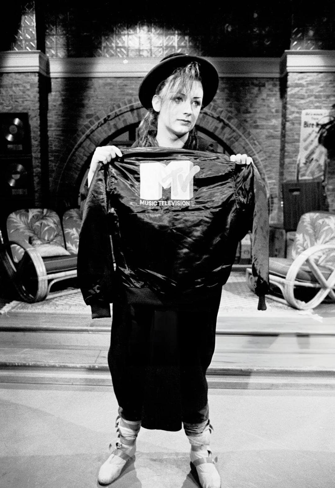 A person wearing a hat and boots holds up a jacket with the MTV Music Television logo, standing in a studio setting with chairs and brick walls in the background. The photo is in black and white.
