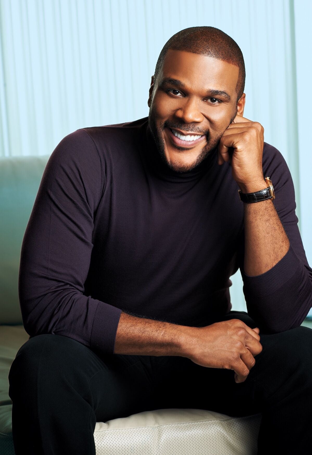 A man with short hair and a beard, wearing a dark turtleneck and watch, sits on a light-colored sofa, smiling and resting his head on his hand.