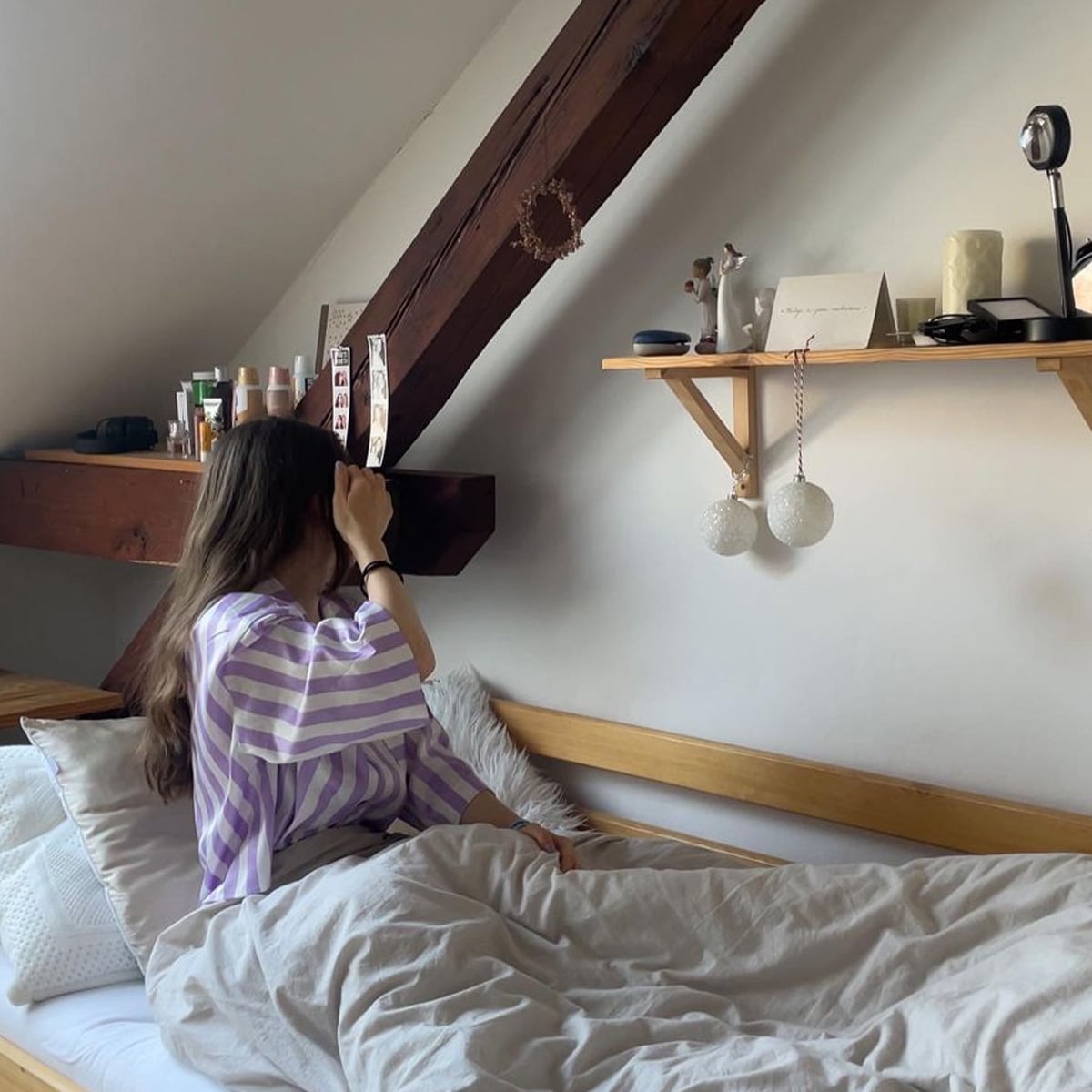 A person with long hair sits on a bed with white and gray bedding, facing away from the camera. They wear a purple and white striped shirt. A wooden shelf with decor and beauty products is mounted on the white wall above.