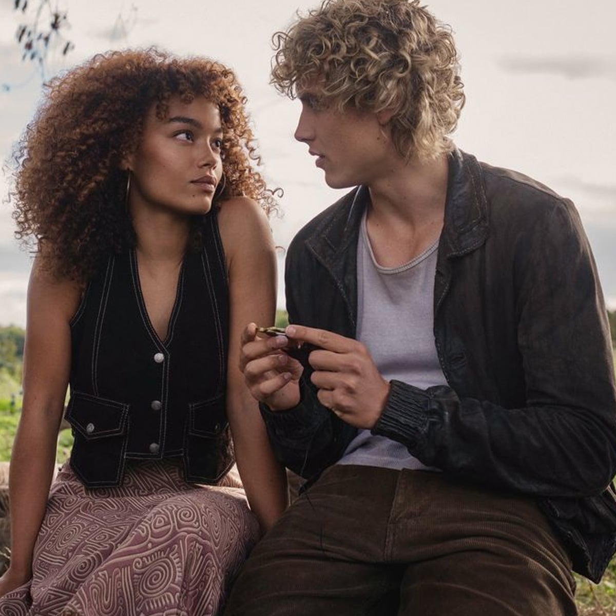 A young woman with curly hair and a patterned skirt sits beside a young man with blond curls and a dark jacket, outdoors. They face each other, appearing mid-conversation, with a cloudy sky in the background.