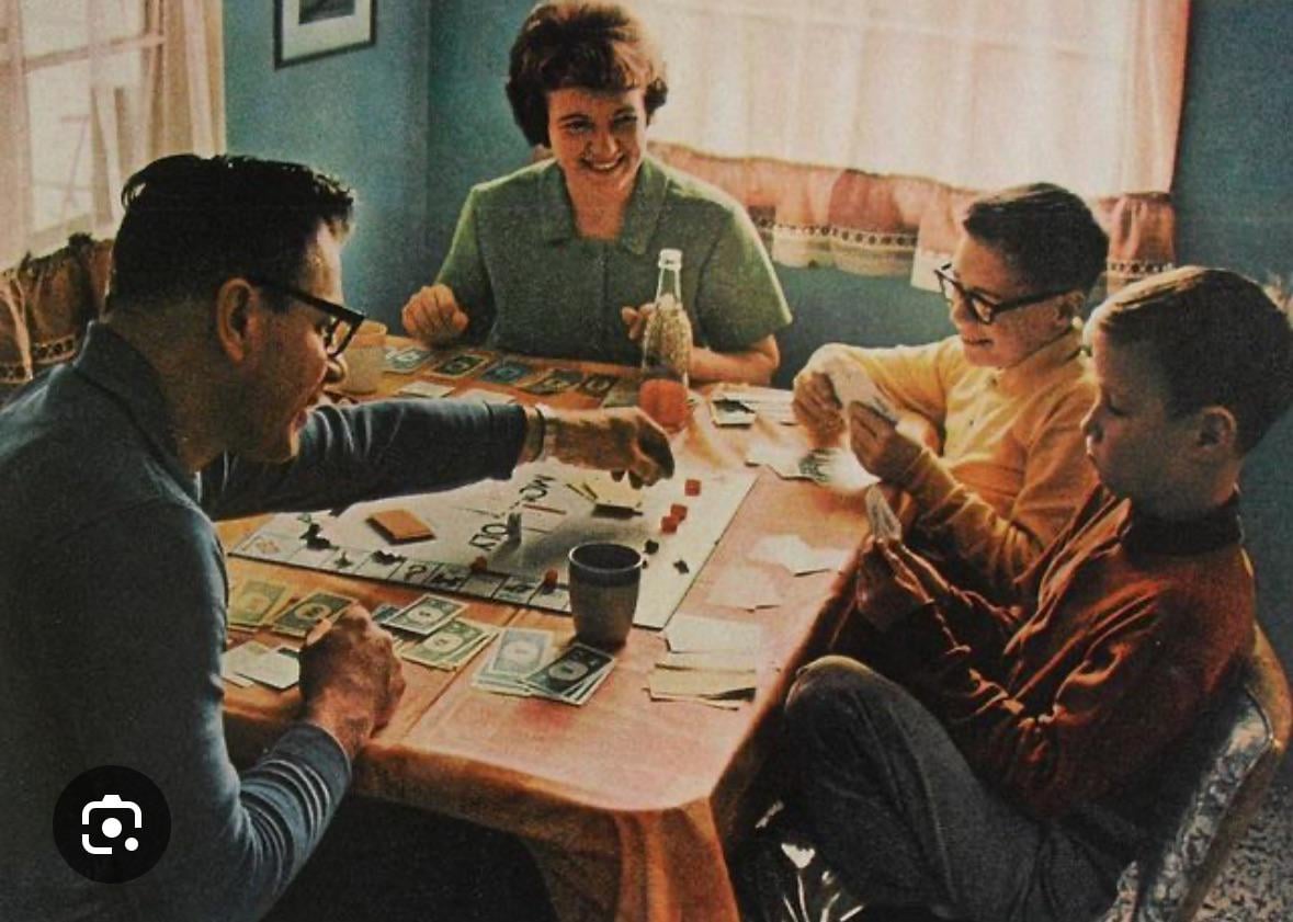 A family of four sits around a table playing Monopoly. The father moves a game piece, the mother smiles, and two young boys focus on their cards. Monopoly money and game pieces are spread out on the table.