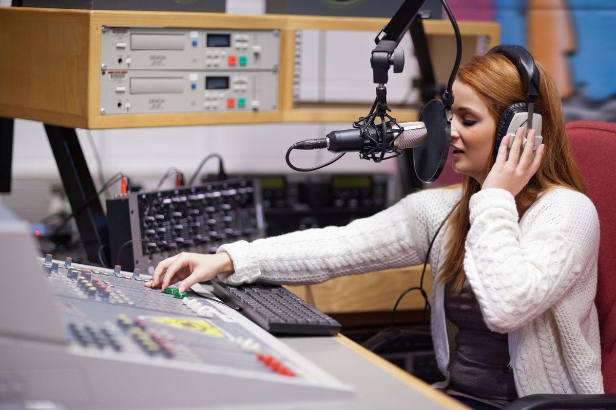 A woman wearing headphones speaks into a microphone while sitting at a radio station control panel, adjusting audio levels and operating broadcasting equipment.
