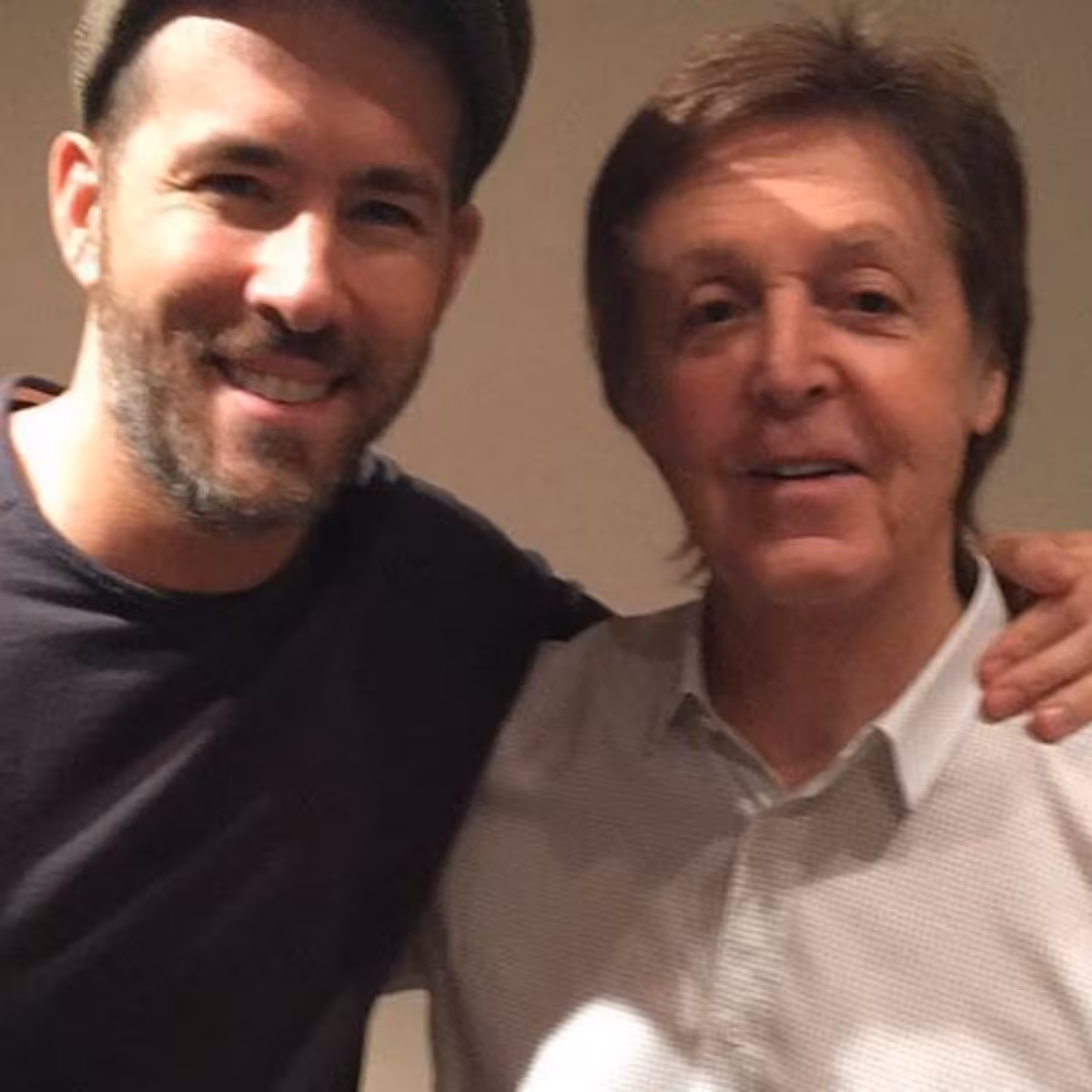 Two men are standing close together and smiling at the camera. One is wearing a flat cap and a dark shirt, while the other is in a light-colored, collared shirt. The man on the left has his arm around the other person.