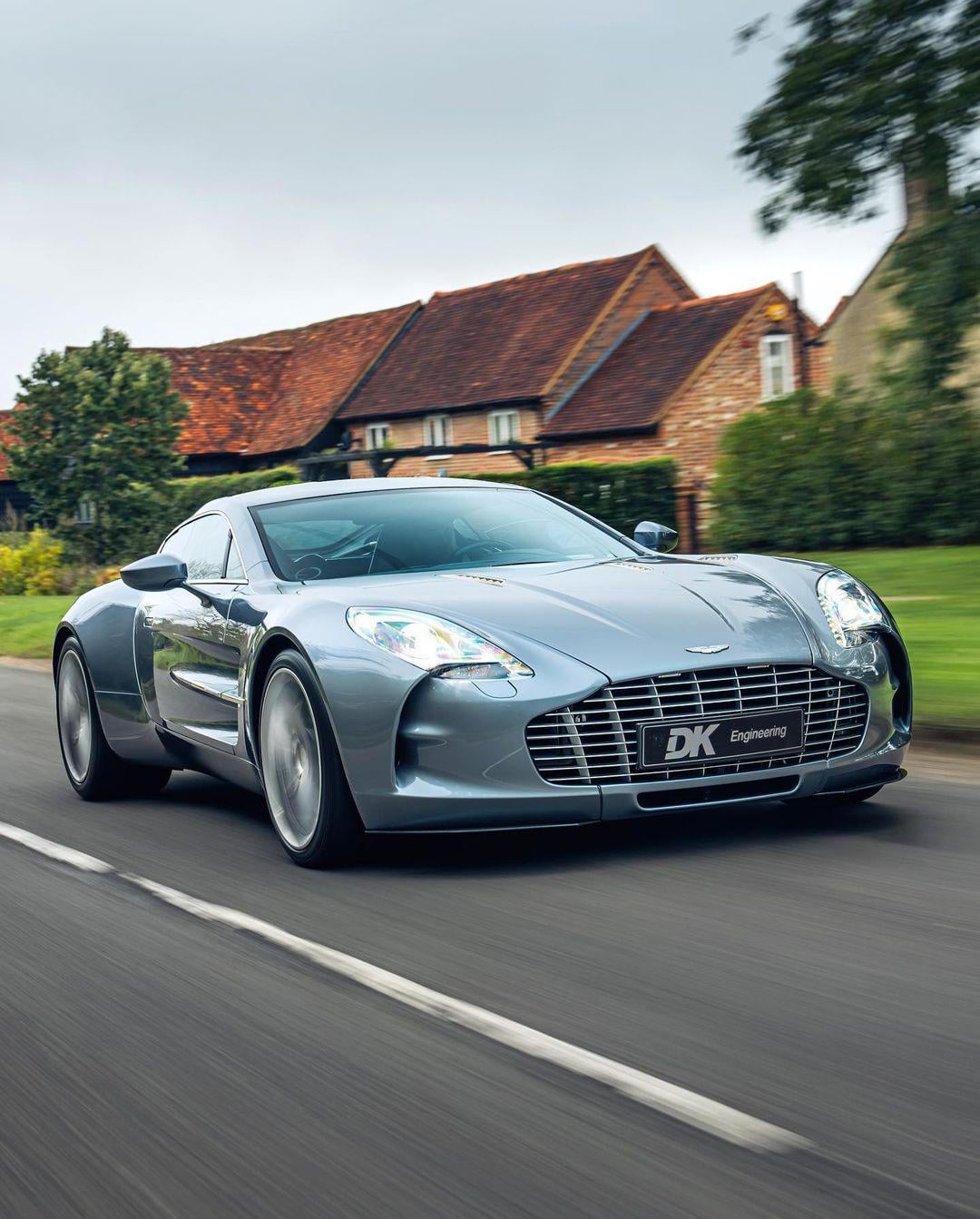 A silver Aston Martin sports car drives on a suburban road, with houses and greenery in the background. The car's sleek design and aerodynamic lines are prominent.