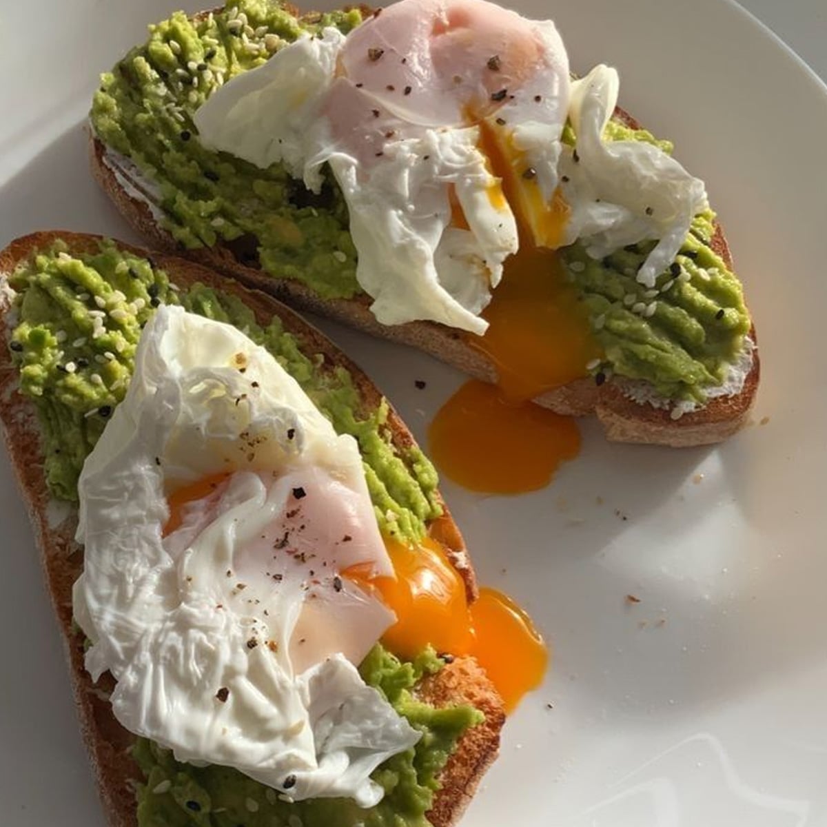 Two slices of toast topped with mashed avocado and poached eggs, with runny yolk spilling onto the plate. The dish is sprinkled with black pepper and served on a white plate.