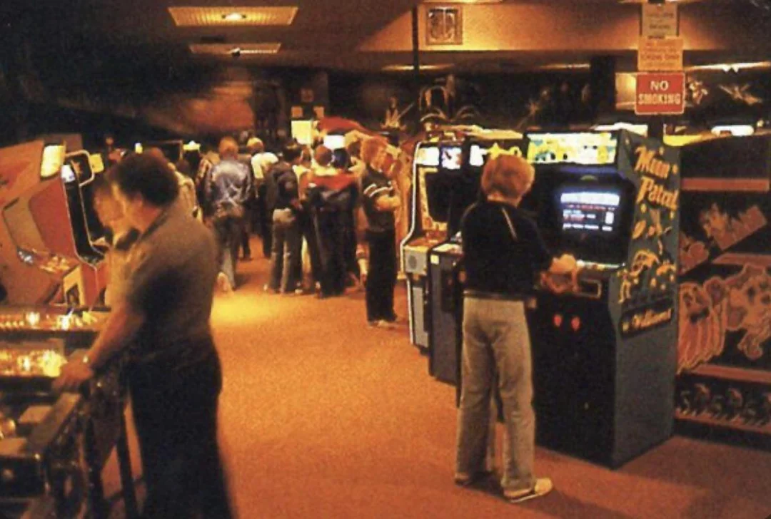 A group of people, mostly teens, play arcade and pinball machines in a dimly lit, crowded retro game arcade. Bright screens and colorful cabinets line the carpeted room.