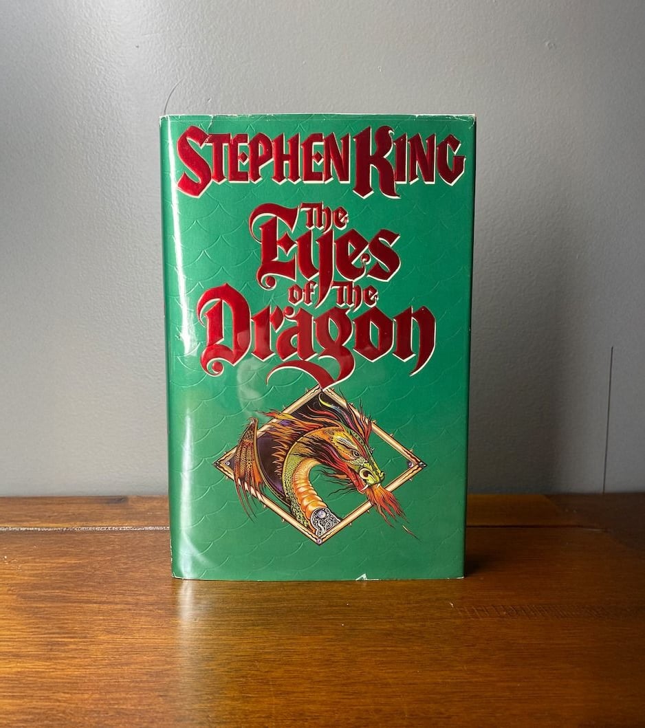 A hardcover copy of "The Eyes of the Dragon" by Stephen King stands upright on a wooden surface. The green cover features bold red and white lettering and an illustrated dragon breathing fire inside a diamond-shaped frame.