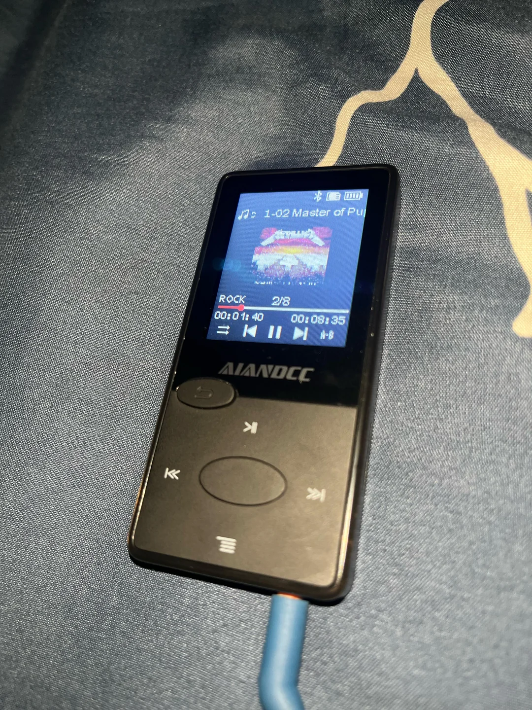 A black MP3 player with a color screen displays the song "1-02 Master of Pu..." in the Rock genre. The device is on a blue surface, and blue earphones are plugged in. Album art is visible on the screen.