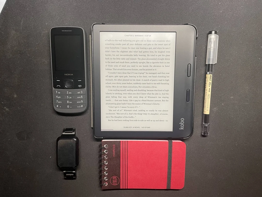 A flat lay photo displays a black Nokia phone, a Kobo e-reader showing text, a black pen, a black smart band, and a small red spiral notepad arranged neatly on a dark surface.