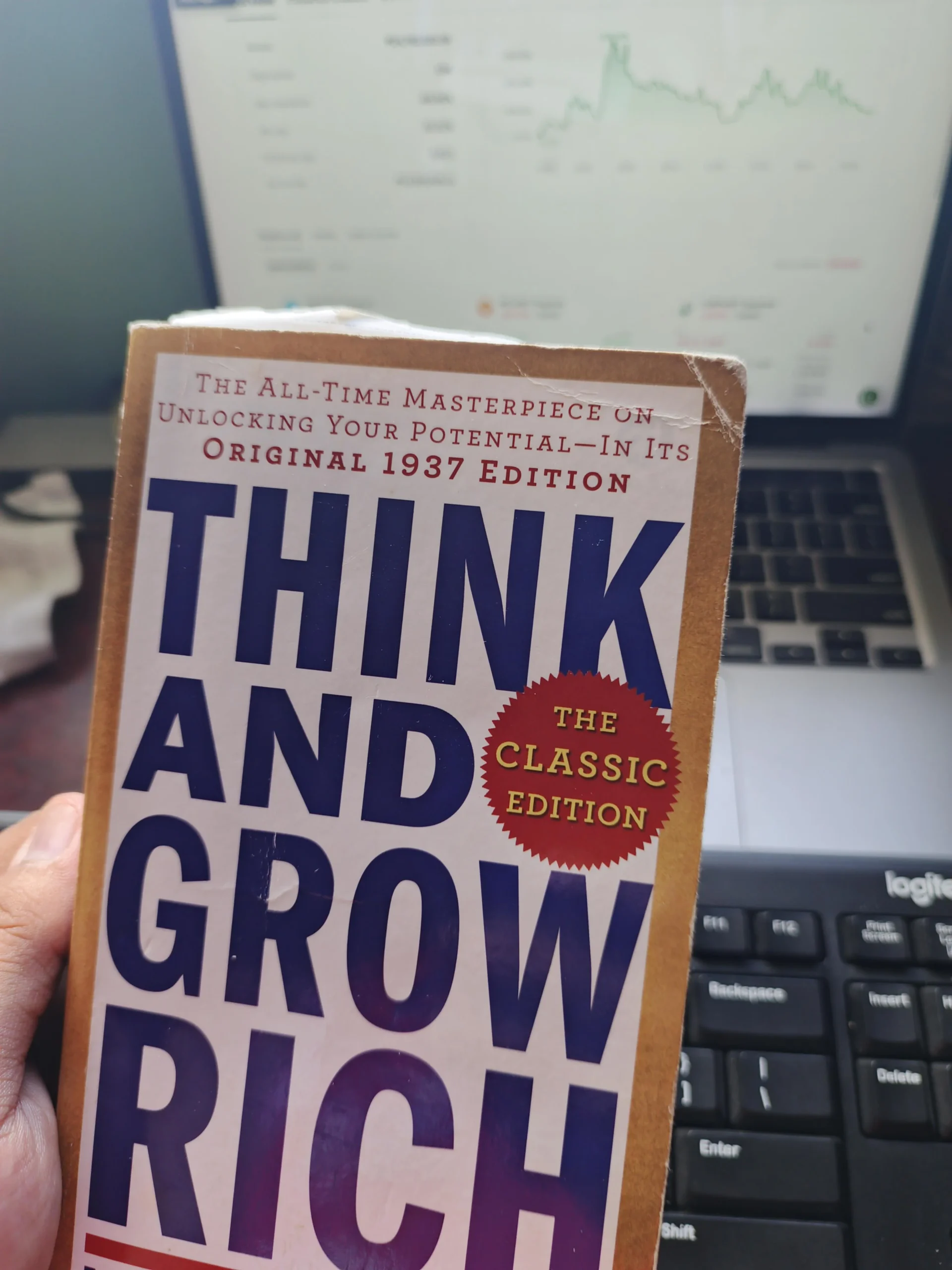 A hand holds a copy of "Think and Grow Rich: The Classic Edition." In the background, there is a laptop with a graph on the screen and part of a keyboard visible.