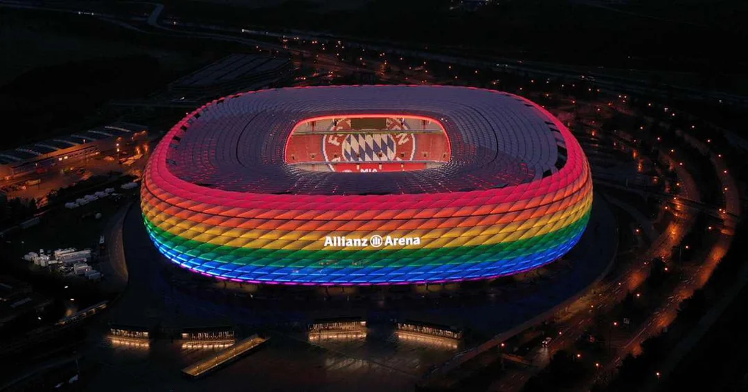 Allianz Arena stadium illuminated with rainbow colors at night, with the Bayern Munich logo displayed on a screen inside the stadium.