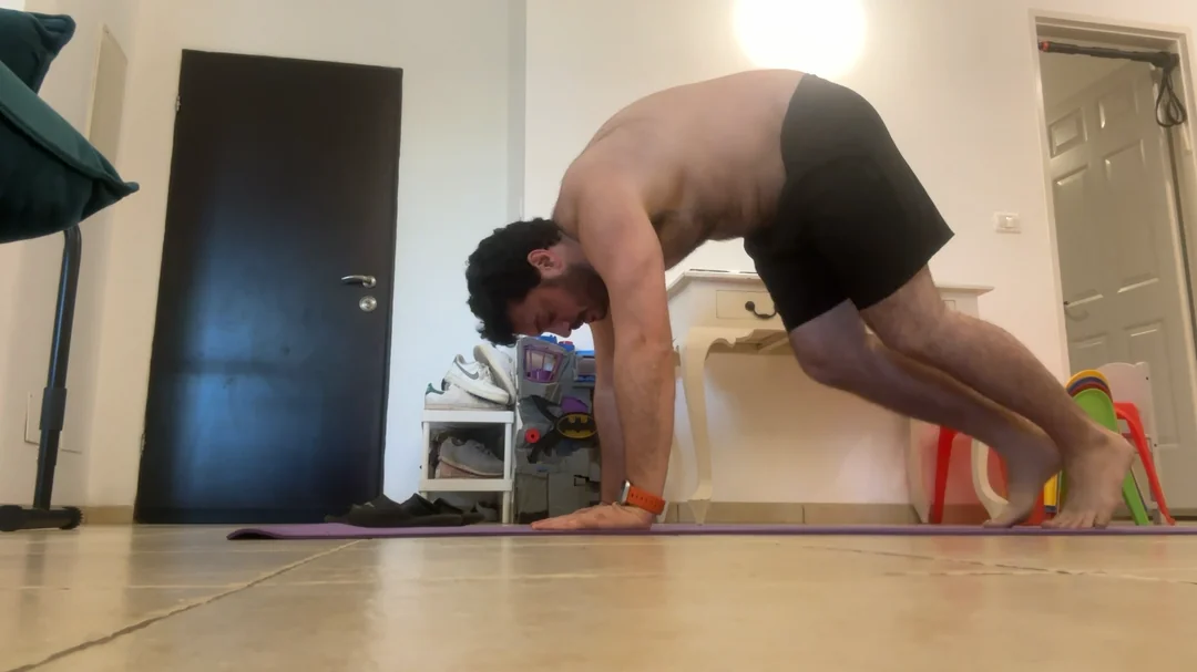 A man in black shorts performs a plank variation on a yoga mat indoors, balancing on his hands and toes with knees bent. The room features a closed door, furniture, and children's toys in the background.