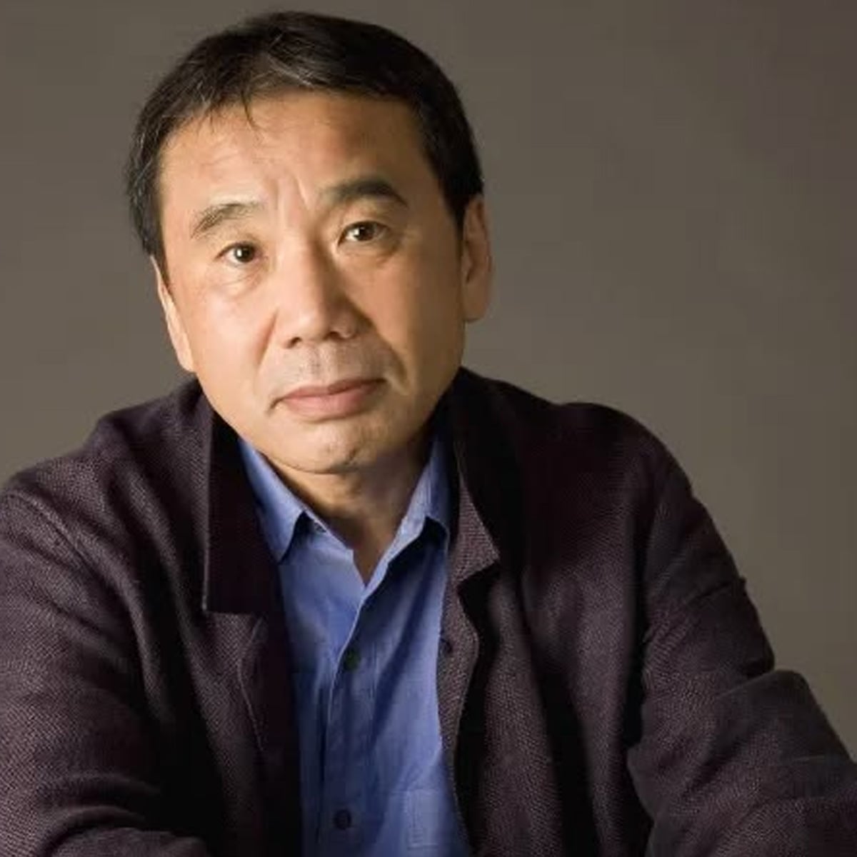A middle-aged person with short dark hair, wearing a blue shirt and dark jacket, gazes thoughtfully at the camera against a plain, neutral background.