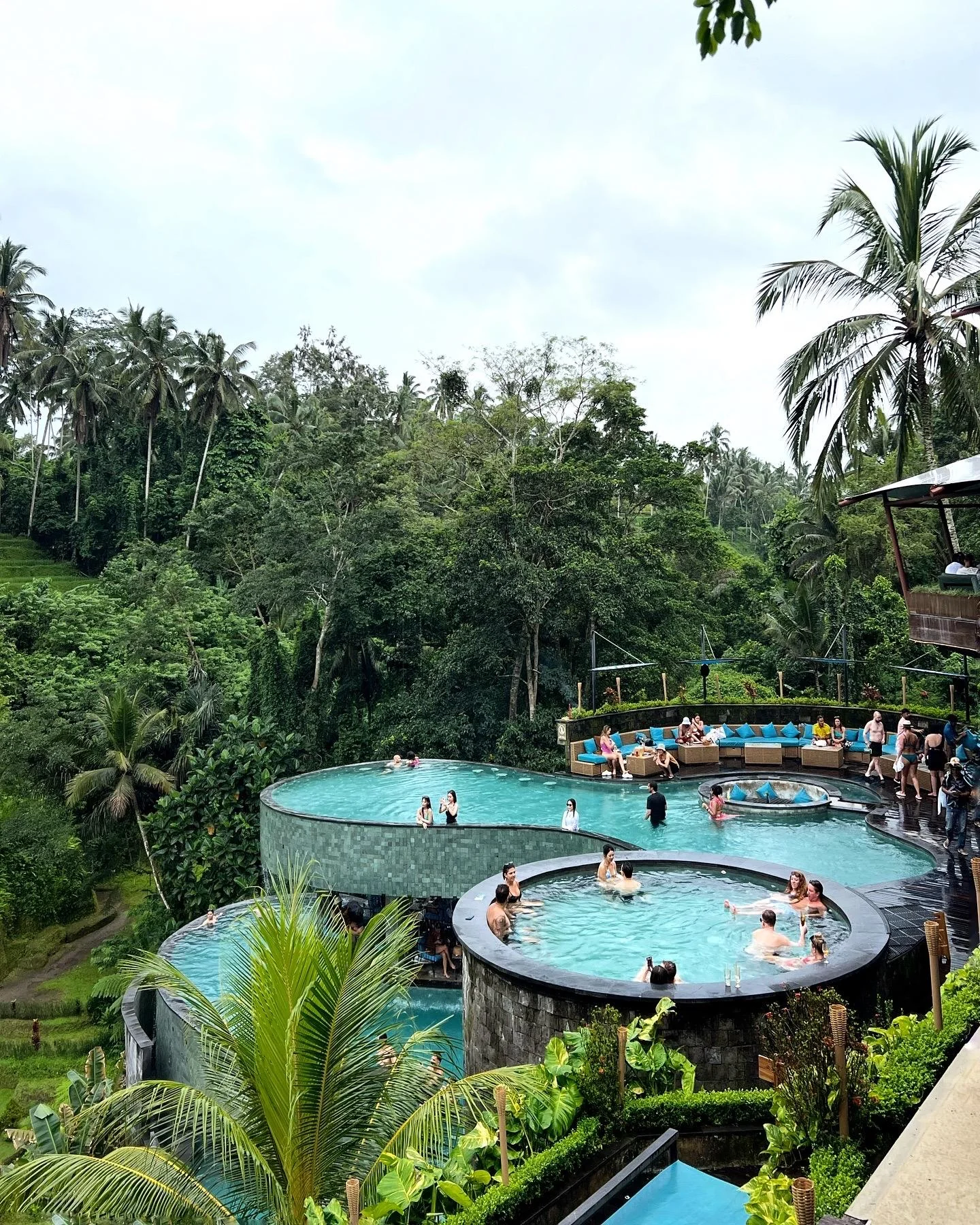 Three circular, tiered infinity pools overlook lush tropical greenery, with people relaxing in the water and on lounge chairs. Palm trees and dense forest create a serene, natural backdrop under an overcast sky.