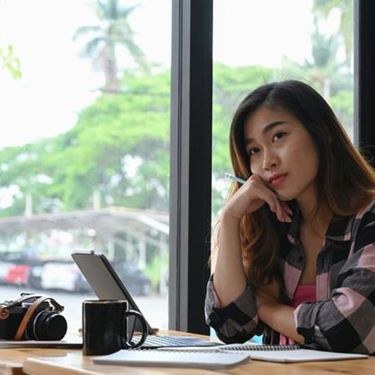 A woman sits at a table by a window, holding a pen and looking thoughtful. In front of her are a tablet, a notebook, a coffee mug, and a camera. Trees and parked cars are visible outside.