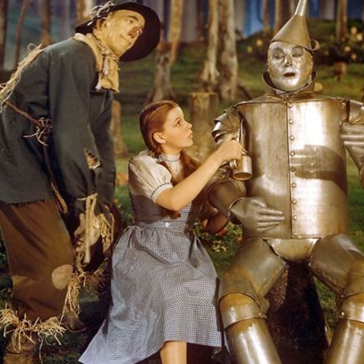 A young girl in a blue gingham dress oils the arm of a tin man while a scarecrow stands beside them in a forest setting, recreating a classic scene from "The Wizard of Oz.