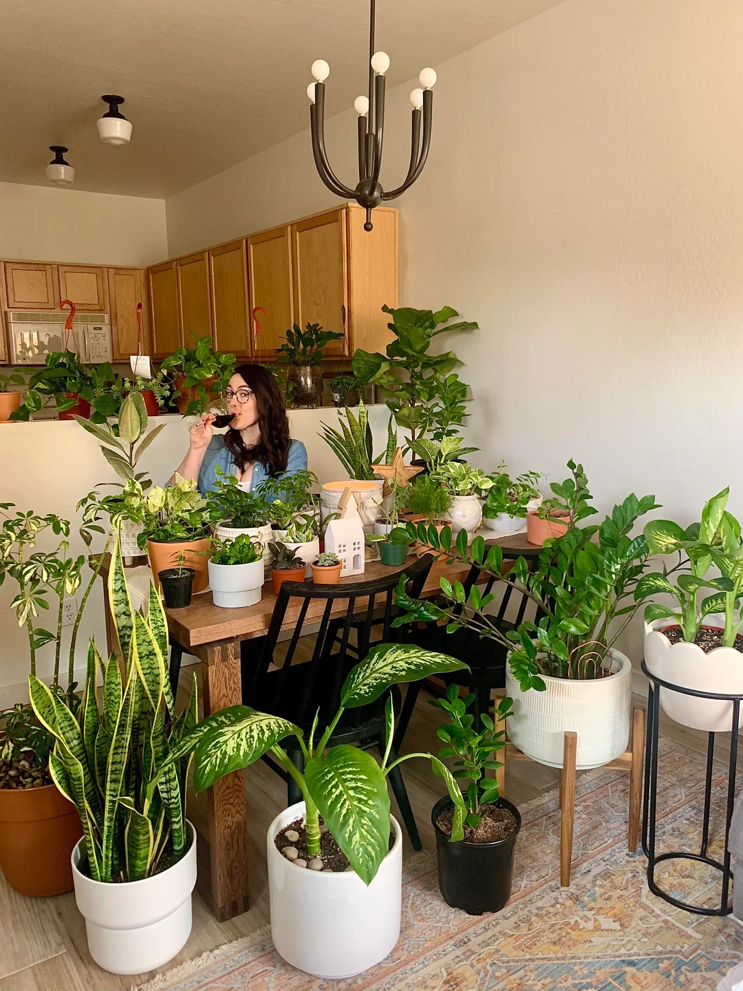 A woman sits at a dining table surrounded by many potted houseplants in a bright room with wooden cabinets, sipping from a mug and smiling. The plants fill the table and floor, creating a lush indoor garden atmosphere.