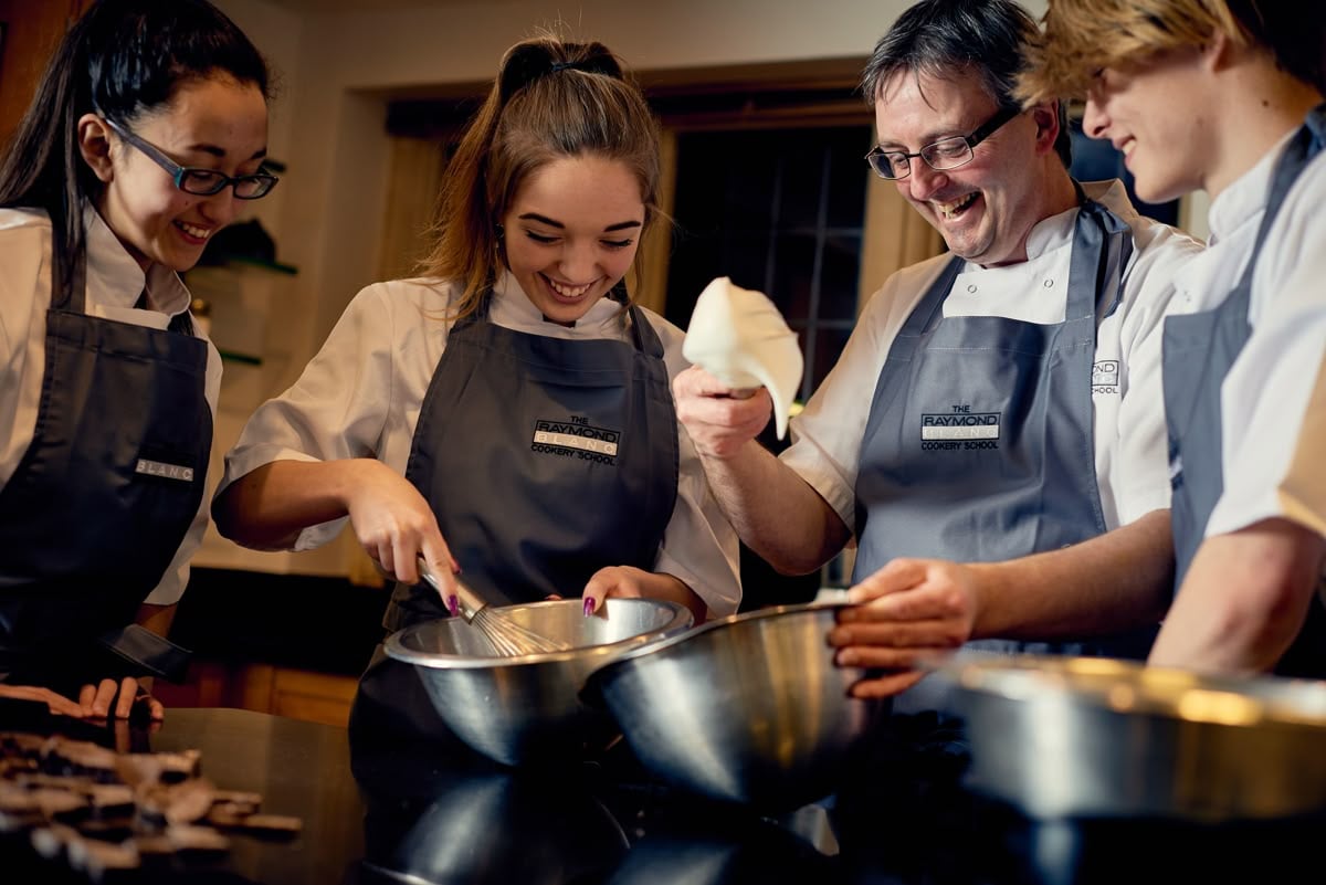 Four people in aprons smile and work together in a kitchen, mixing ingredients in metal bowls; one is piping cream while others watch and participate, suggesting a cooking class or group baking activity.