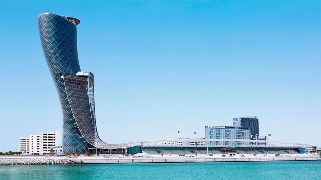 A futuristic, leaning skyscraper with a unique twisted design stands beside modern low-rise buildings on a waterfront under a clear blue sky.